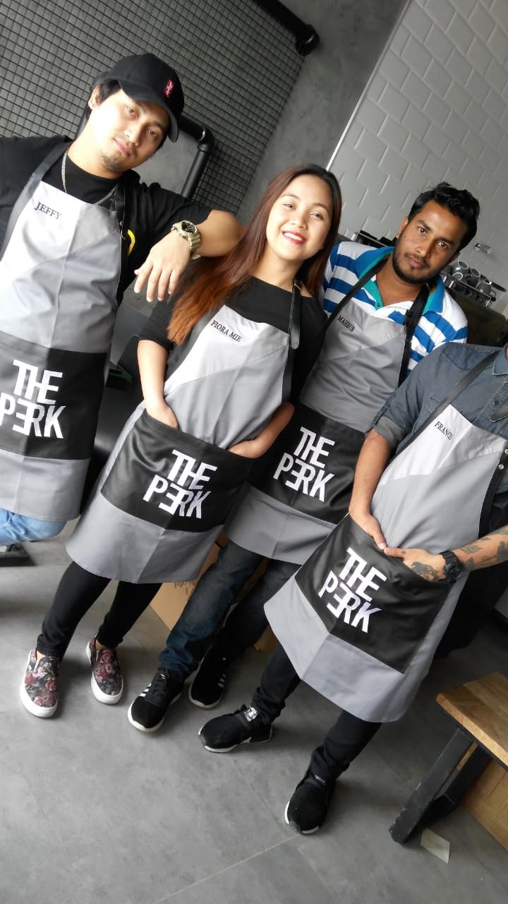 Four café staff posing together in matching grey-and-black bib aprons printed 'The Perk'