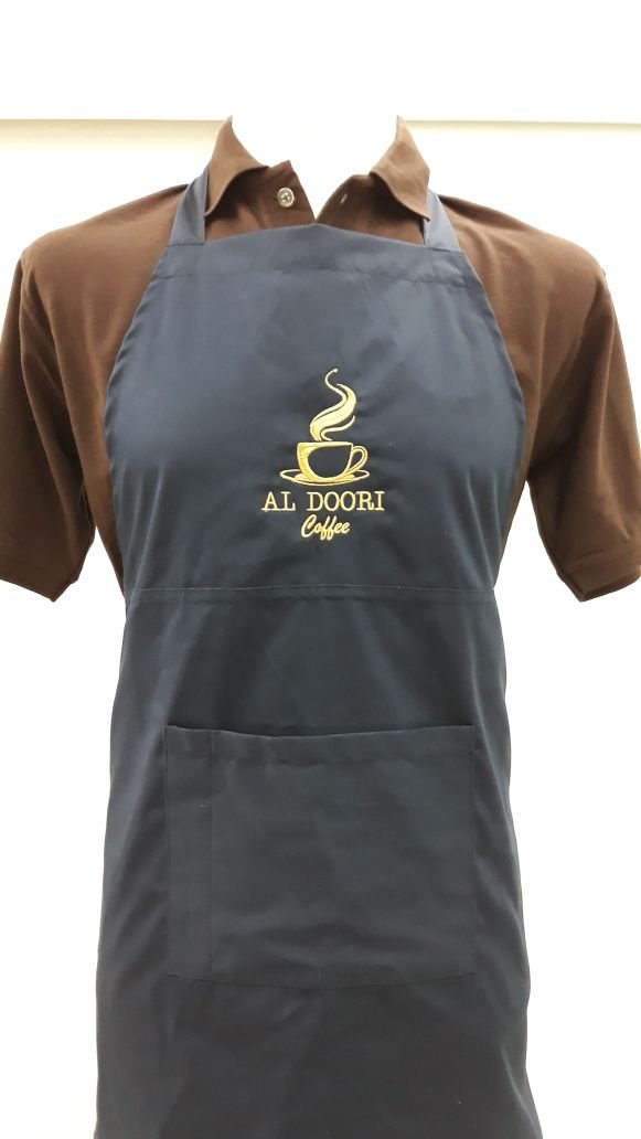 Grey bib apron with gold embroidered 'Al Doori Coffee' logo worn over a brown shirt
