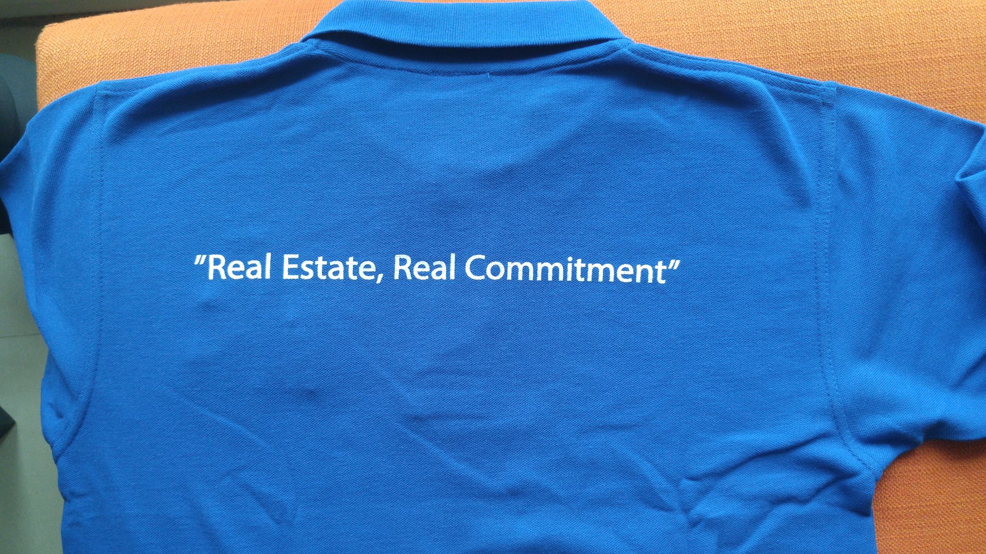 Back of a royal-blue polo shirt with the white tagline 'Real Estate, Real Commitment'