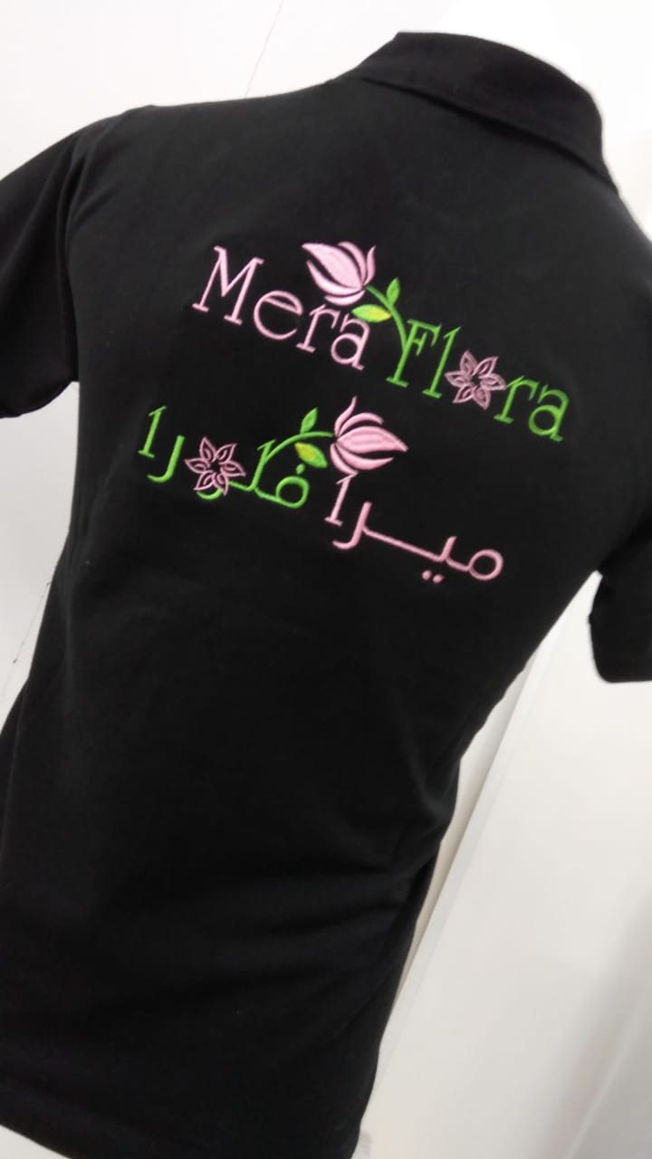 Back of a black polo shirt with a pink-and-green embroidered floral logo in English and Arabic lettering